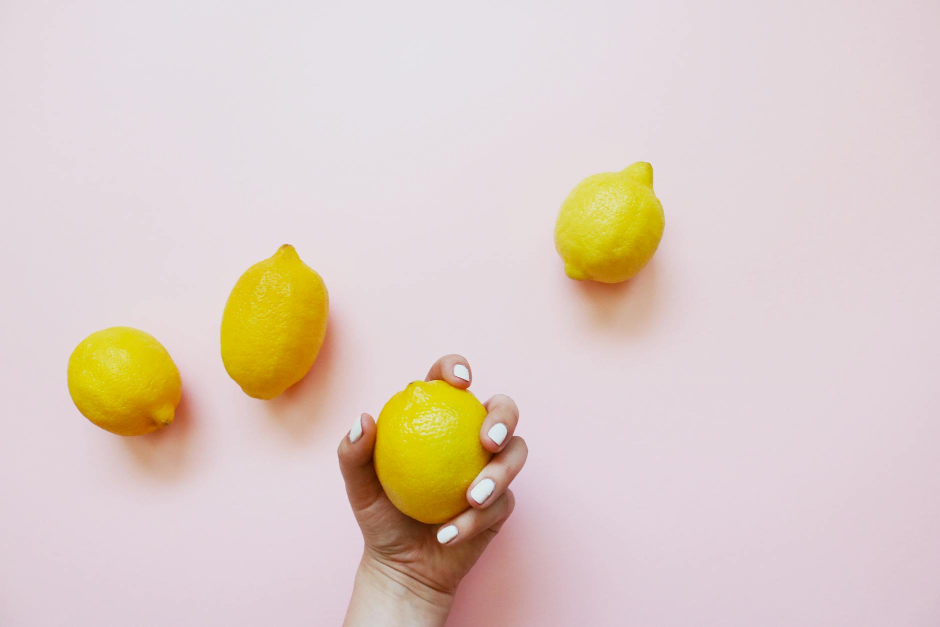 A hand holding a fresh lemon on a soft pink background, surrounded by three additional lemons.