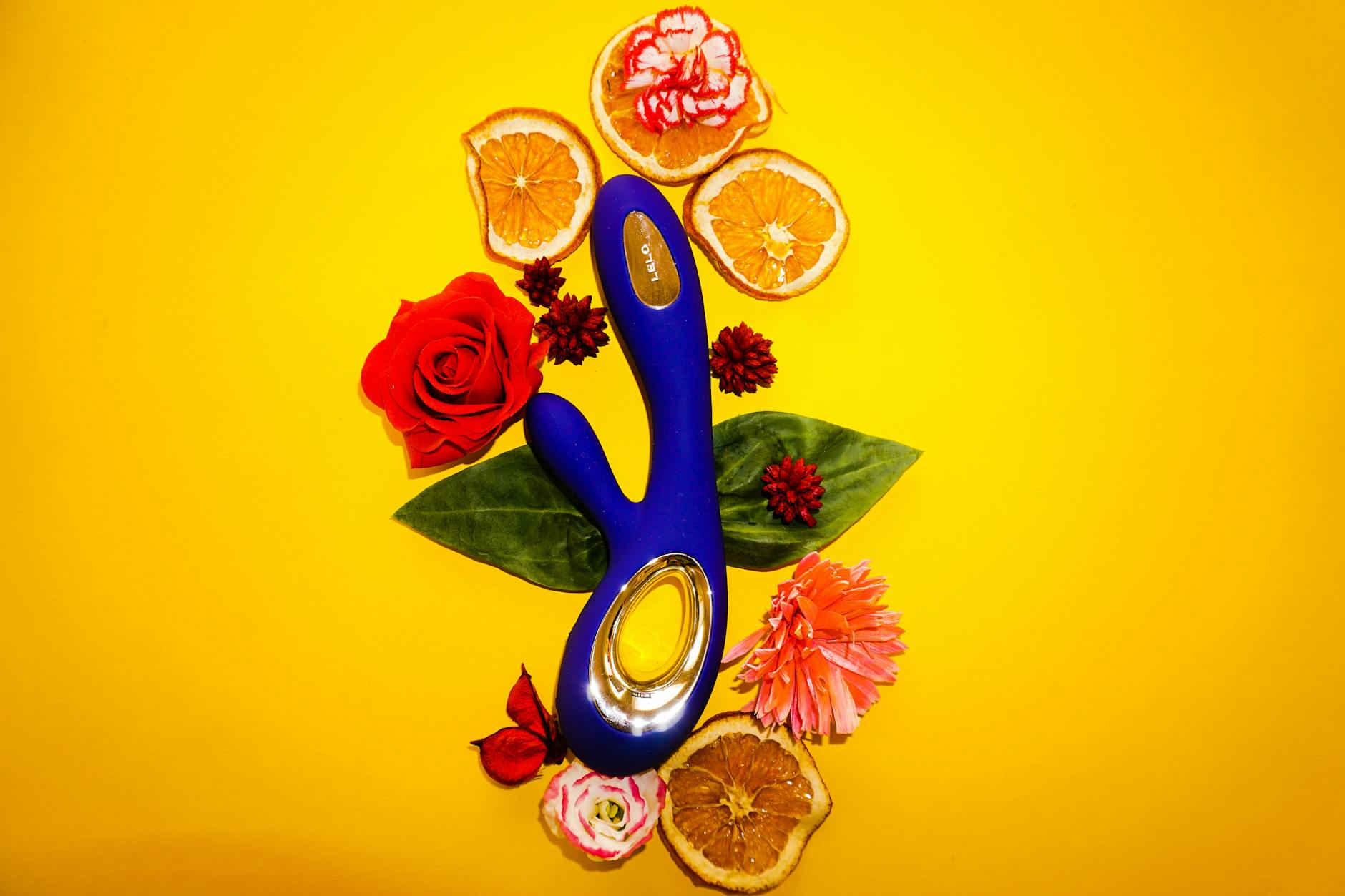 Vibromasseur clitoral bleu entouré de fleurs et de tranches d'orange sur fond jaune